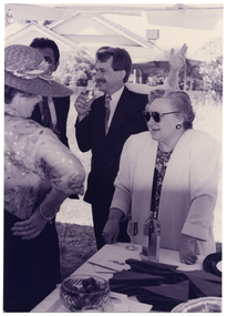 B&W medium close up photo of a few people gathered under a marquee, outdoors. A man is drinking from a champagne flute. Behind him, someone (partially obscured) is raising one arm. In the foreground, a woman is leaning on the table while chatting to another woman wearing a straw hat. On the table, there is a bottle of wine, a flute champagne glass, a bowl of strawberries, stacks of paper napkins, and a few brochures. In the background, there is a house and some trees.