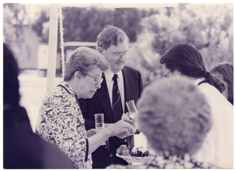B&W medium close up photo of a few people gathered under a marquee, outdoors. A waitress (partially obscured) presents a fruit and cheese platter to two people. A woman chooses something from the tray while a man looks on. He is holding a champagne flute and a small sandwich in one hand. A few other people (partially obscured) are evident in the foreground and background.