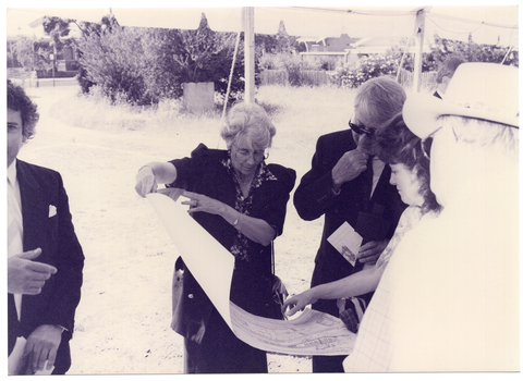 B&W medium close up photo of a few people gathered under a marquee, outdoors. Three people are looking at a large piece of paper while a fourth person (at the edge of the right side of the frame) looks on. Another person (partially obscured) is standing to the left edge of the frame.