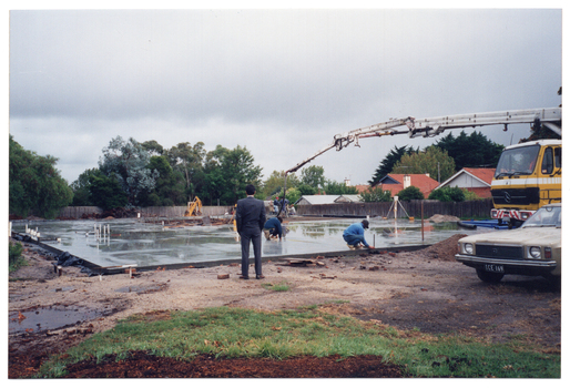 COL photo of the crane of a truck hovering over one section of a large concrete slab on a construction site. A man in a suit observes while construction workers attend to various sections of the slab. In front of the truck, there is a brown car with the registration number plate ICE-169.