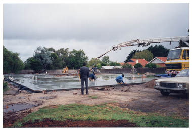 COL photo of the crane of a truck hovering over one section of a large concrete slab on a construction site. A man in a suit observes while construction workers attend to various sections of the slab. In front of the truck, there is a brown car with the registration number plate ICE-169.