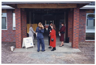 COL photo of five people standing at the entrance of a newly built reddish-brown brick building. At the front of the entrance, on the left, there is a "Move it or Lose it" A-frame sandwich board next to a brick pillar. Behind it, there is a board with a closed gold curtain resting on an easel or A-framed stand. At the back of the entrance, to the right, there is a "Move it or Lose it" vertical sign or banner (partially obscured).