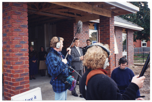 COL photo of a man standing near a microphone on a stand. Behind him are two other men (partially obscured). They are at the front entrance of a newly built reddish-brown brick building. In the background, there are a few people are standing at the rear of the front entrance. In the foreground, there are three media sound crew. One is holding up a boom microphone on long pole. Another is holding another long pole. A third is sitting or crouching down. They are all wearing headphones. In the bottom left corner of the frame, there is a "Move it or Lose it" A-frame sandwich board (partially obscured).