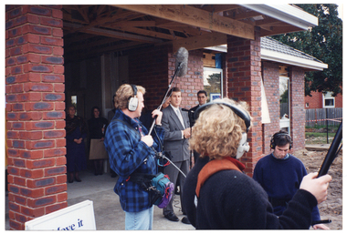 COL photo of a man standing near a microphone on a stand. Behind him are two other men (partially obscured). They are at the front entrance of a newly built reddish-brown brick building. In the background, there are a few people are standing at the rear of the front entrance. In the foreground, there are three media sound crew. One is holding up a boom microphone on long pole. Another is holding another long pole. A third is sitting or crouching down. They are all wearing headphones. In the bottom left corner of the frame, there is a "Move it or Lose it" A-frame sandwich board (partially obscured).