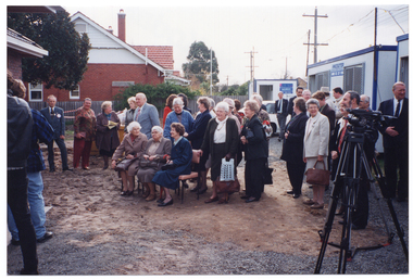 COL photo of a group of people gathered outside a brick building. Three elderly women are seated on three chairs, while the rest of the crowd are standing behind and beside them. The ground below consists of bare earth and gravel. In the foreground, to the right of the frame, there is a television or video camera on a tripod. In the background, to the right of the frame, there are three portable construction site offices. There is a red brick house on the property next door, behind a wooden fence.