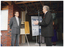 COL photo of two men standing at the front entrance of a reddish-brown brick building. One of them is behind a microphone on a stand. Behind them, there is an easel onto which rests a rectangular bronze plaque on a board. The board has a small gold curtain, which has been drawn open to reveal the plaque. There is also a freestanding, vertical "Move it or Lose it" banner on a stand. There are two people (partially obscured) standing in the background.