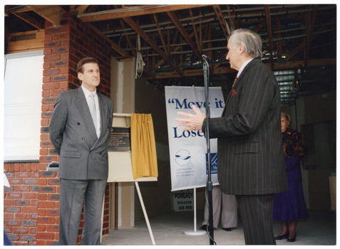COL photo of two men standing at the front entrance of a reddish-brown brick building. One of them is behind a microphone on a stand. Behind them, there is an easel onto which rests a rectangular bronze plaque on a board. The board has a small gold curtain, which has been drawn open to reveal the plaque. There is also a freestanding, vertical "Move it or Lose it" banner on a stand. There are two people (partially obscured) standing in the background.