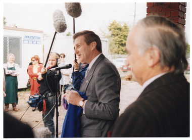 COL photo of two men standing in the front entrance of a building. One of the men is holding a piece of bright blue fabric. Beyond them, there are two people (one partially obscured) each holding a long pole. On the end of each pole, there is a microphone with a fluffy grey cover. They are poised above the man behind the microphone. In the background, there are a few people standing in front of two portable construction site offices.