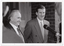 B&W close up photo of two men standing in front of a brick building, smiling broadly. In the foreground, there is a microphone on a stand (partially obscured).