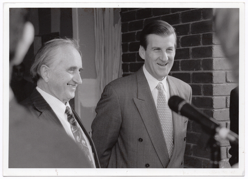 B&W close up photo of two men standing in front of a brick building, smiling broadly. In the foreground, there is a microphone on a stand (partially obscured).