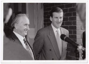 B&W close up photo of two men standing in front of a brick building, smiling broadly. In the foreground, there is a microphone on a stand (partially obscured).