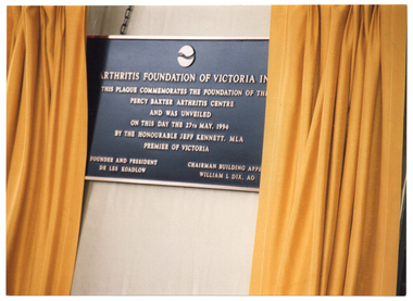 COL close up photo of a bronze plaque hanging from a chain on a board. The board has two gold curtains which have been drawn open to reveal the plaque. The text on the plaque is: "ARTHRITIS FOUNDATION OF VICTORIA IN... [partially obscured] THIS PLAQUE COMMEMORATES THE FOUNDATION OF TH... [partially obscured] PERCY BAXTER ARTHRITIS CENTRE AND WAS UNVEILED ON THIS DAY THE 27TH MAY, 1994 BY THE HONOURABLE JEFF KENNETT, MLA PREMIER OF VICTORIA FOUNDER AND PRESIDENT DR LES KOADLOW CHAIRMAN BUILDING APPE... [partially obscured] WILLIAM L DIX, AO".