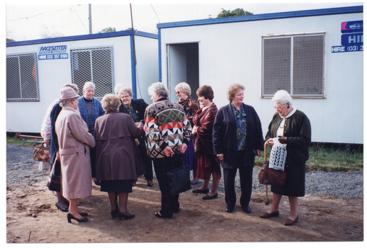 COL photo of a small group of women standing in front of two portable construction site offices, chatting. One woman is holding a wooden walking stick with a curved handle. The ground below consists of bare earth and gravel.