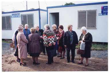COL photo of a small group of women standing in front of two portable construction site offices, chatting. One woman is holding a wooden walking stick with a curved handle. The ground below consists of bare earth and gravel.