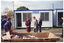 COL photo of a few people on a construction site. Several people (two partially obscured) are walking past some portable site offices. In the foreground, a man sits on the edge of a skip bin full of discarded building materials and cardboard boxes, while another man is standing behind him. They are both looking at the people walking past.