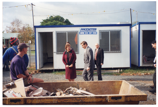 COL photo of a few people on a construction site. Several people (two partially obscured) are walking past some portable site offices. In the foreground, a man sits on the edge of a skip bin full of discarded building materials and cardboard boxes, while another man is standing behind him. They are both looking at the people walking past.