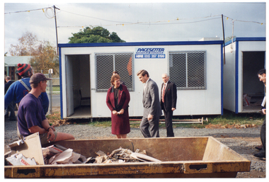 COL photo of a few people on a construction site. Several people (two partially obscured) are walking past some portable site offices. In the foreground, a man sits on the edge of a skip bin full of discarded building materials and cardboard boxes, while another man is standing behind him. They are both looking at the people walking past.