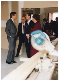 COL photo of four people chatting inside an unfinished new building. In the foreground, there is a circular saw poised over a plank of timber. In the background, there are several people walking through the other rooms in the building.