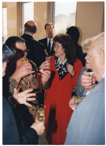 COL photo of a small group of people standing close to one another in a circle. They are each holding a champagne flute, filled with champagne and/or orange juice. In the background, there are several other people also standing close together.