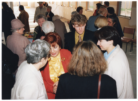 COL photo of a group of people standing close to one another, chatting. In the background, a round table covered with a tablecloth is evident in a gap between some people. There are platters of food on the table. The photo has been taken from a high angle, highlighting a small group of people in the foreground.