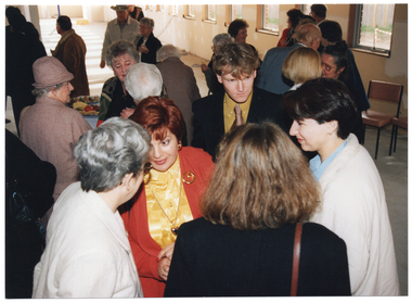 COL photo of a group of people standing close to one another, chatting. In the background, a round table covered with a tablecloth is evident in a gap between some people. There are platters of food on the table. The photo has been taken from a high angle, highlighting a small group of people in the foreground.