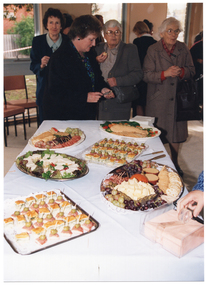 COL photo of a group of people standing close to one another at the end of a table. Some of them are eating or have some food in their hands. On the table in front of them, there are several platters of finger food and two stacks of paper napkins.  In the foreground, one person's hand is reaching for some paper napkins in a clear plastic napkin holder. There are several other people standing in the background, and some chairs near a large window.