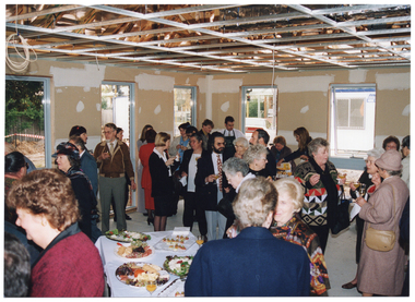 COL photo of a big group of people standing inside a large room in an unfinished building. Several people are holding glasses of wine or champagne in one hand. In the foreground, there is a table containing several large platters of finger food. Above them, the roof trusses and electrical wiring are evident as a ceiling has not yet been installed. The walls have not yet been painted. Large windows reveal views of an unlandscaped garden and a portable construction site office on the outside.