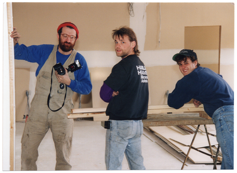 COL photo of three men inside an unfinished building. One of them is leaning on some planks of timber which are resting across a long trestle table. He is smiling. The other two men are standing near him. They are all looking directly at the camera.