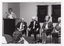 B&W photo of a woman standing behind a lectern, speaking into a microphone. Beside her, there are four people seated on a row of chairs. Hanging on the wall behind them are two "Move It or Lose It" banners. In the foreground, one person (partially obscured) has been Photographed from behind.