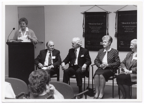 B&W photo of a woman standing behind a lectern, speaking into a microphone. Beside her, there are four people seated on a row of chairs. Hanging on the wall behind them are two "Move It or Lose It" banners. In the foreground, one person (partially obscured) has been Photographed from behind.
