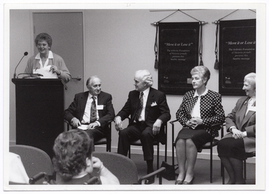 B&W photo of a woman standing behind a lectern, speaking into a microphone. Beside her, there are four people seated on a row of chairs. Hanging on the wall behind them are two "Move It or Lose It" banners. In the foreground, one person (partially obscured) has been Photographed from behind.