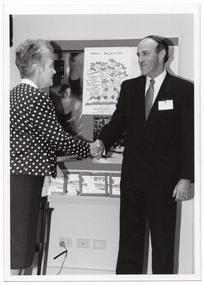 B&W photo of a woman and a man shaking hands. Behind them is a display board with various posters, including an illustrated poster titled, "Step out for strong bones". On a small table beneath the display board, there are several small framed illustrations of a building leaning against the wall. The woman appears to have two framed items in her other hand.