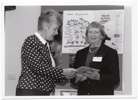 B&W photo of a woman presenting a small framed item to another woman. The woman receiving the item is looking directly at the camera. Behind them is a display board with various posters, including an illustrated poster titled "Step out for strong bones". There is a small framed picture (partially obscured) beneath the display board.