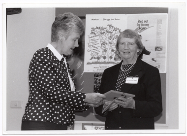 B&W photo of a woman presenting a small framed item to another woman. The woman receiving the item is looking directly at the camera. Behind them is a display board with various posters, including an illustrated poster titled "Step out for strong bones". There is a small framed picture (partially obscured) beneath the display board.