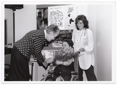 B&W photo of three women. One woman (who is a wheelchair user) is holding a floral bouquet. Another woman is leaning forward and shaking her hand. A third woman is standing behind the wheelchair. Behind the three women is a display board with various posters, including two illustrated posters - one titled "Arthritis - you can feel better", and the other titled "Step out for strong bones". A third poster has the title (partially obscured), "... more mileage ...of your day". On a small table beneath the display board, there are several small framed illustrations of a building leaning against the wall.