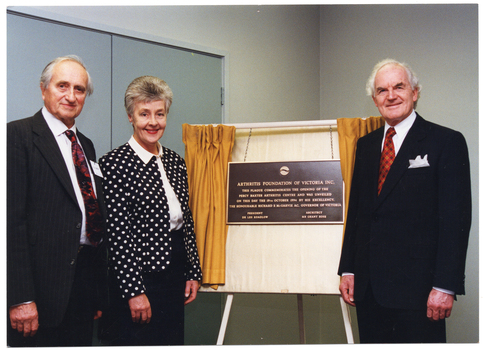 COL photo of three people standing beside a plaque, which is handing on a board resting on an easel. Across the board is a small set of gold curtains, which have been drawn open to reveal the plaque. The text on the plaque is: "ARTHRITIS FOUNDATION OF VICTORIA INC. THIS PLAQUE COMMEMORATES THE OPENING OF THE PERCY BAXTER ARTHRITIS CENTRE AND WAS UNVEILED ON THIS DAY THE 19TH OCTOBER 1994 BY HIS EXCELLENCY, THE HONOURABLE RICHARD E McGARVIE AC, GOVERNOR OF VICTORIA PRESIDENT DR LES KOADLOW ARCHITECT MR GRANT ROSE".