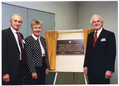 COL photo of three people standing beside a plaque, which is handing on a board resting on an easel. Across the board is a small set of gold curtains, which have been drawn open to reveal the plaque. The text on the plaque is: "ARTHRITIS FOUNDATION OF VICTORIA INC. THIS PLAQUE COMMEMORATES THE OPENING OF THE PERCY BAXTER ARTHRITIS CENTRE AND WAS UNVEILED ON THIS DAY THE 19TH OCTOBER 1994 BY HIS EXCELLENCY, THE HONOURABLE RICHARD E McGARVIE AC, GOVERNOR OF VICTORIA PRESIDENT DR LES KOADLOW ARCHITECT MR GRANT ROSE".