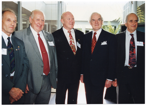 COL photo of five men, standing in a row next to one another, posing for a photo inside a room. Behind them is an open door leading to a courtyard. There are some large fern fronds evident in the courtyard. On either side of the open doorway, there are wide windows with thin venetian blinds.