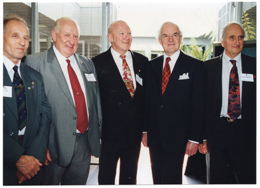 COL photo of five men, standing in a row next to one another, posing for a photo inside a room. Behind them is an open door leading to a courtyard. There are some large fern fronds evident in the courtyard. On either side of the open doorway, there are wide windows with thin venetian blinds.