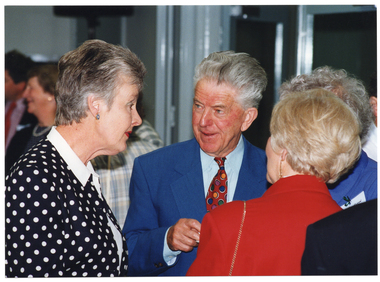 COL close up photo of four people, standing and chatting to one another in a crowded room. One woman has been Photographed from behind, and another person is partially obscured. There are other people standing in the background.