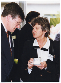 COL close up photo of two people standing in a crowded room, chatting. One of them is holding a cup and saucer.