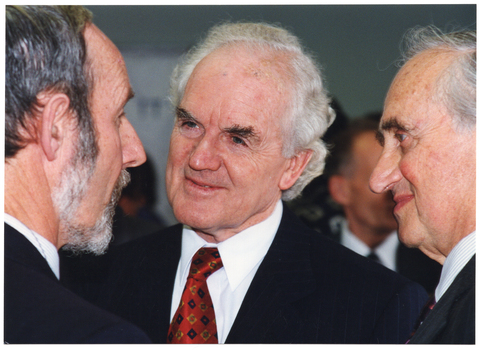 COL close up photo of three men standing in a crowded room, chatting.