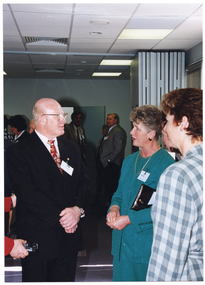 COL close up photo of three people chatting inside a crowded room. In the background, there are several other people standing and chatting.