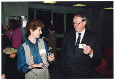 COL medium close up photo of a woman and a man chatting inside a crowded room. In the background, there are several other people standing and chatting.