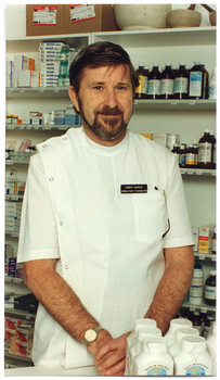COL photo of a man standing behind a pharmacy counter. There are some medications on the counter in front of him, and a wall of shelves full of various medications behind him.