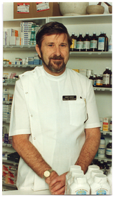 COL photo of a man standing behind a pharmacy counter. There are some medications on the counter in front of him, and a wall of shelves full of various medications behind him.