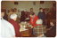 COL photo of a large group of people gathered around a room. In the centre, there is a wooden table with various cakes, biscuits, and two sugar bowls on it. One woman is cutting a cake. In the background, there is a freestanding display board with posters on it and various framed pictures on the wall. There are also two electric heaters on the wall.