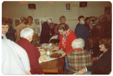 COL photo of a large group of people gathered around a room. In the centre, there is a wooden table with various cakes, biscuits, and two sugar bowls on it. One woman is cutting a cake. In the background, there is a freestanding display board with posters on it and various framed pictures on the wall. There are also two electric heaters on the wall.
