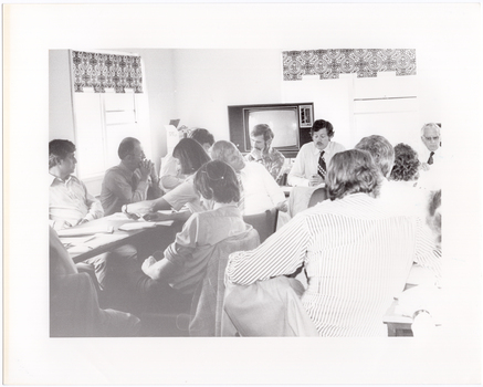 B&W photo of a group of people sitting around some tables placed in a large square formation. Some people are sitting on the inside of the square table formation. There are several documents on every table.  In the background, there is a large television set on a stand, with an open box on top of a small table next to it. There are also two windows with blinds.