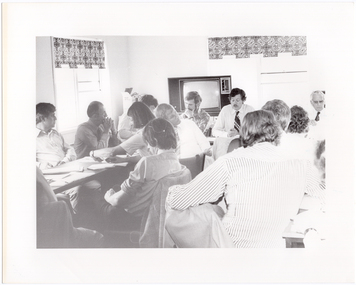 B&W photo of a group of people sitting around some tables placed in a large square formation. Some people are sitting on the inside of the square table formation. There are several documents on every table.  In the background, there is a large television set on a stand, with an open box on top of a small table next to it. There are also two windows with blinds.